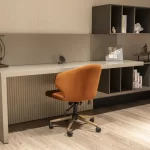 Modern genç odası çalışma köşesi; krem rengi masa, turuncu kadife sandalye ve duvara monte raf sistemi.